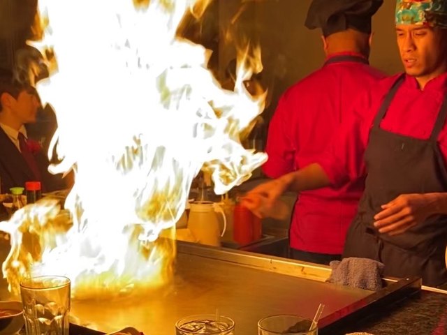 A Hakata chef in a red jacket sends a tall column of flame up from the hibachi grill as guests watch.