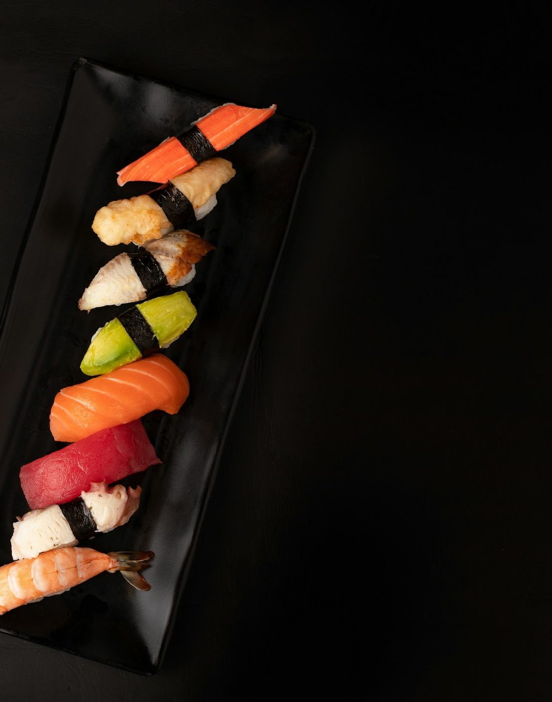 Nigiri lined up on a black slate beside chopsticks and soy sauce.