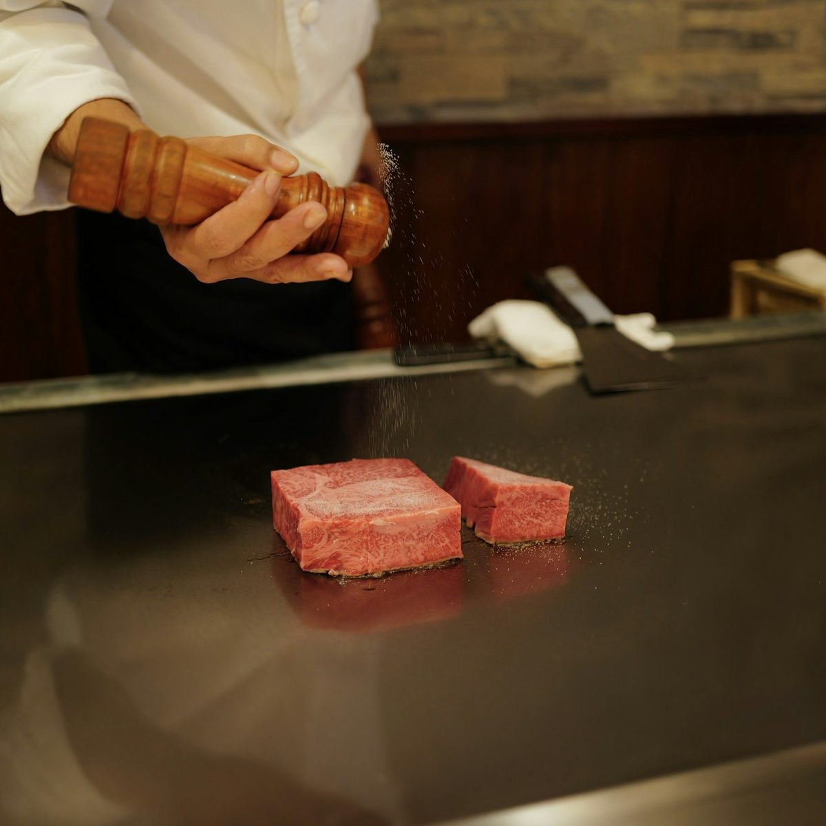 A chef's knife beside two blocks of marbled beef on a polished steel teppan grill.