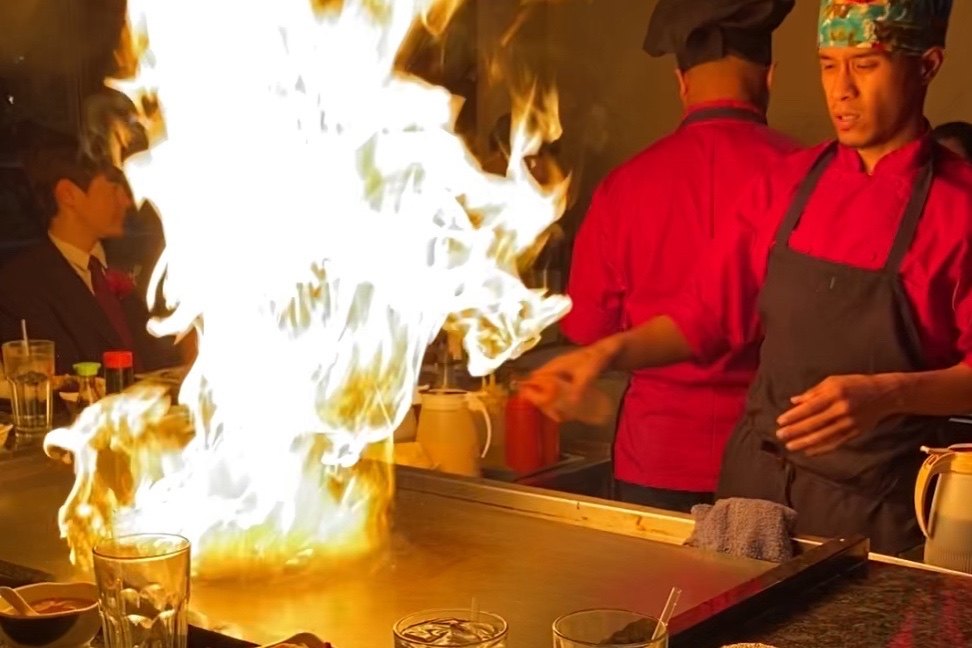 Hakata chefs in red jackets at the hibachi grill as a column of flame rises in front of guests.